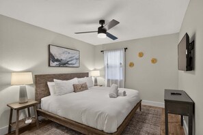 A hotel room with a bed, bedside lamps, a ceiling fan, a painting, and a television.