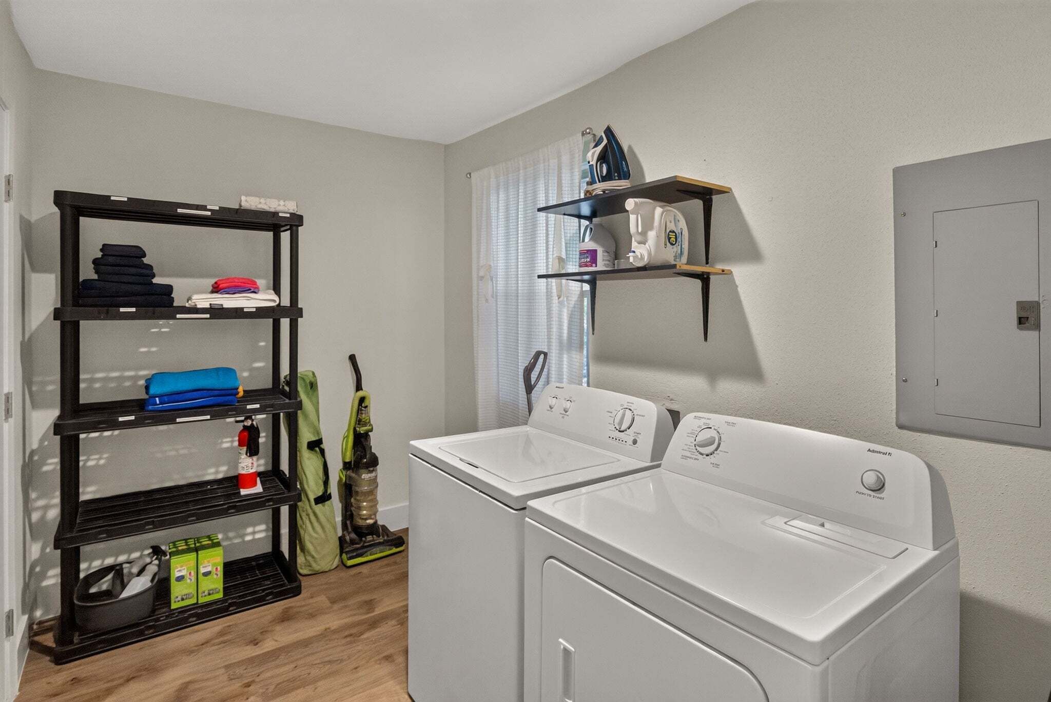 A laundry room with two white washing machines, a black shelving unit with folded clothes, and a window with blinds.