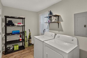 A laundry room with two white washing machines, a black shelving unit with folded clothes, and a window with blinds.