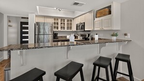 A modern kitchen with white cabinets, a stainless steel refrigerator, a microwave, and a granite countertop. There are three black stools at the island.
