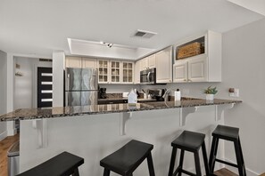 A modern kitchen with white cabinets, a stainless steel refrigerator, a microwave, and a granite countertop. There are three black stools at the island.