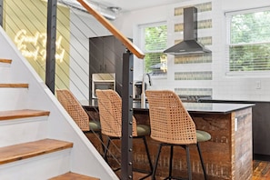 A modern kitchen with a wooden staircase, wicker bar stools, and a neon sign. - Olive & Brick | Hot Tub • Private Backyard Oasis (Cincinnati)