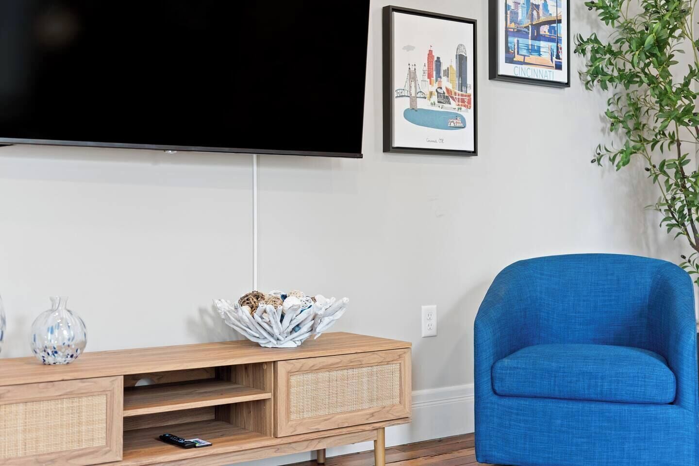 A living room with a flat-screen TV mounted on the wall, a blue armchair, a wooden entertainment unit with a decorative bowl, and framed cityscape pictures.