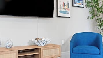 A living room with a flat-screen TV mounted on the wall, a blue armchair, a wooden entertainment unit with a decorative bowl, and framed cityscape pictures.