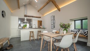A modern kitchen with a central island, wooden stools, and a dining table with chairs. - Luxury Two Bedroom Holiday Cottage with Hot Tub and Sea Views | Y-Ddol (Dyffryn Ardudwy)