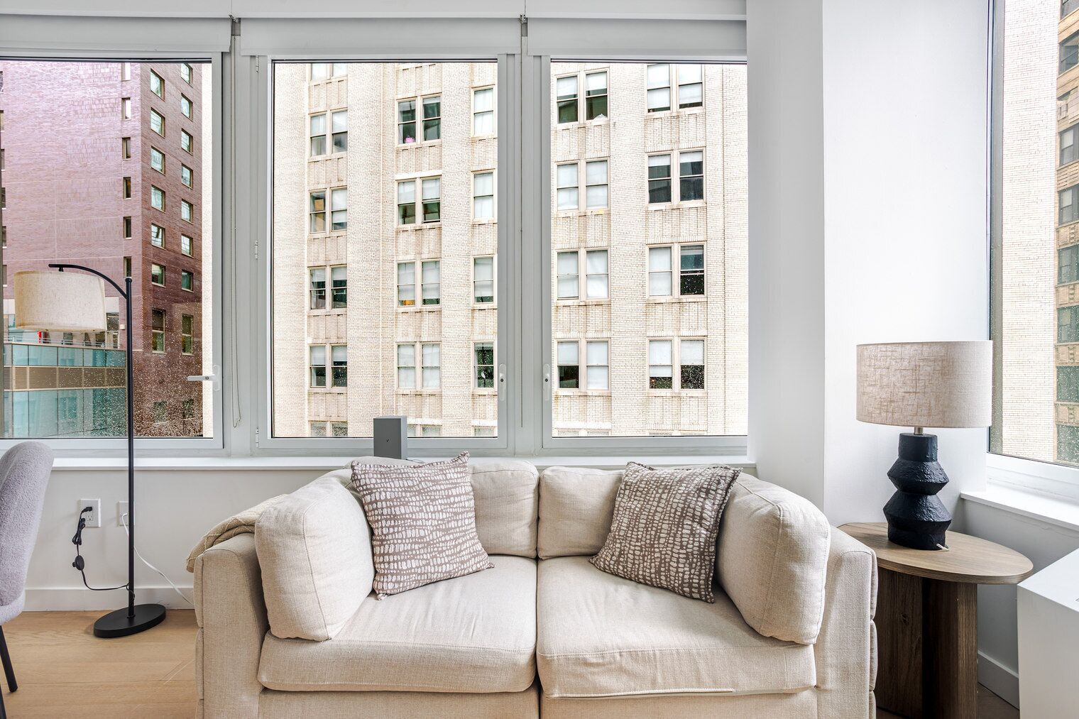 Ein modernes Wohnzimmer mit einem beigefarbenen Sofa, zwei gemusterten Kissen, einem Beistelltisch mit Lampe und Blick auf ein Backsteingebäude durch große Fenster.