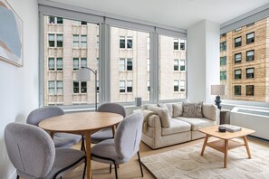 A modern living room with a round wooden table, grey upholstered chairs, a beige sofa, and a small wooden coffee table. Large windows offer views of a brick building. - Blueground | Financial District, rooftop pool, nr Brooklyn Bridge (New York)