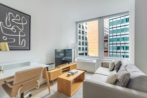 Ein modernes Wohnzimmer mit Flachbildfernseher, weißem Schreibtisch, einer Couch und einem großen Fenster mit Blick auf die Stadt.