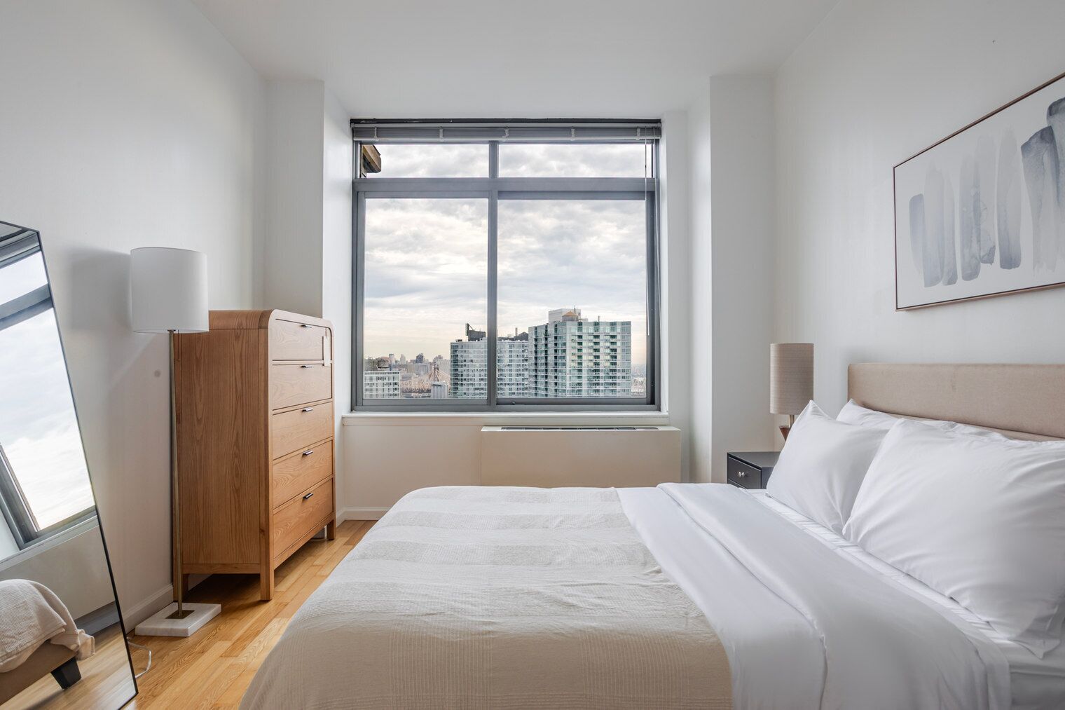 A bedroom with a large window, a bed, a dresser, and a city view.
