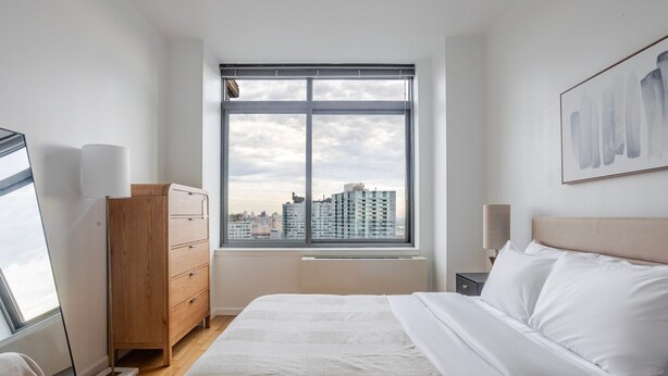 A bedroom with a large window, a bed, a dresser, and a city view.