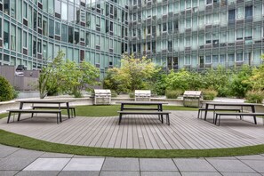 Eine Außenpatio-Terrasse mit Picknicktischen und einem Grill, umgeben von einer Rasenfläche und verschiedenen Pflanzen, vor einem modernen Glasgebäude.