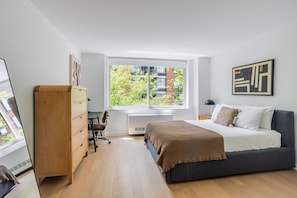 A bedroom with a bed, a wooden dresser, a desk, and a window with a view of buildings and trees. - Blueground | Manhattan, rooftop, nr Battery Park (New York)