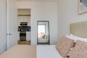 Ein Schlafzimmer mit einem Bett, Kissen, einem Nachttisch und Blick durch eine Tür in einen Küchenbereich mit Geräten.