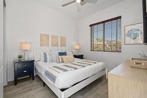 A bedroom with a bed, two bedside tables, a ceiling fan, a window with blinds, and wall art. - NEW Villa Solara by GPM - Swim up bar + Games + More! Walk to festivals, Montage (Indio)
