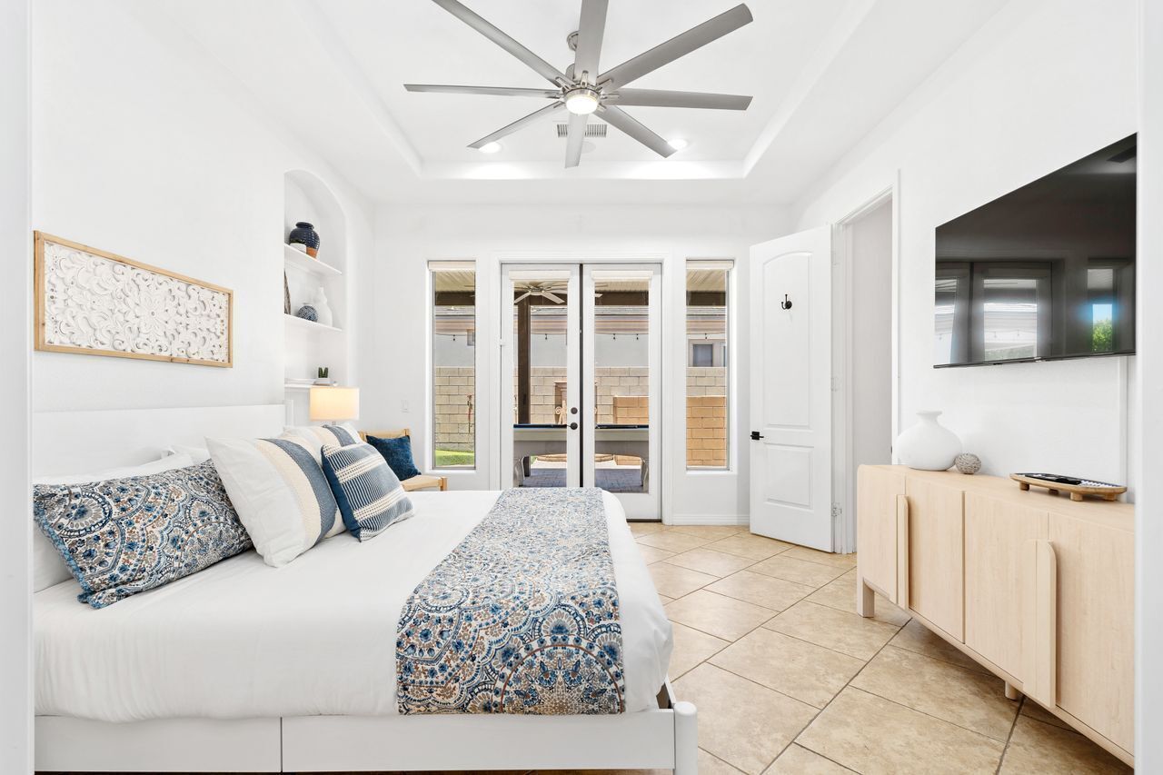 Ein Schlafzimmer mit einem Bett, einem Deckenventilator, einem an der Wand montierten Fernseher und einer Tür, die auf einen Balkon führt.