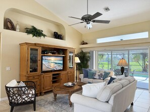 A spacious living room with a television, a ceiling fan, and a view of a garden through large windows. - 3BR Heated Pool Villa - Golf & Beaches (Rotonda West)