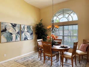 A dining area with a round table, six chairs, abstract wall art, a large window with blinds, and a hanging light fixture. - 3BR Heated Pool Villa - Golf & Beaches (Rotonda West)