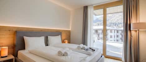 Ein Hotelzimmer mit einem großen Bett, einem Nachttisch mit Lampe und Blick auf eine verschneite Landschaft durch das Fenster.