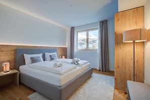 Ein modernes Hotelzimmer mit einem großen Bett, einem Nachttisch mit Lampe, einem Holzschrank und Blick auf schneebedeckte Berge durch das Fenster.