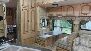 A compact RV interior with a kitchenette, a dining area, and a seating space with a view of the outdoors. - Camper 6 Enjoyable 2-bedroom recreational vehicle with AC just out of Fair Play. (Fair Play)