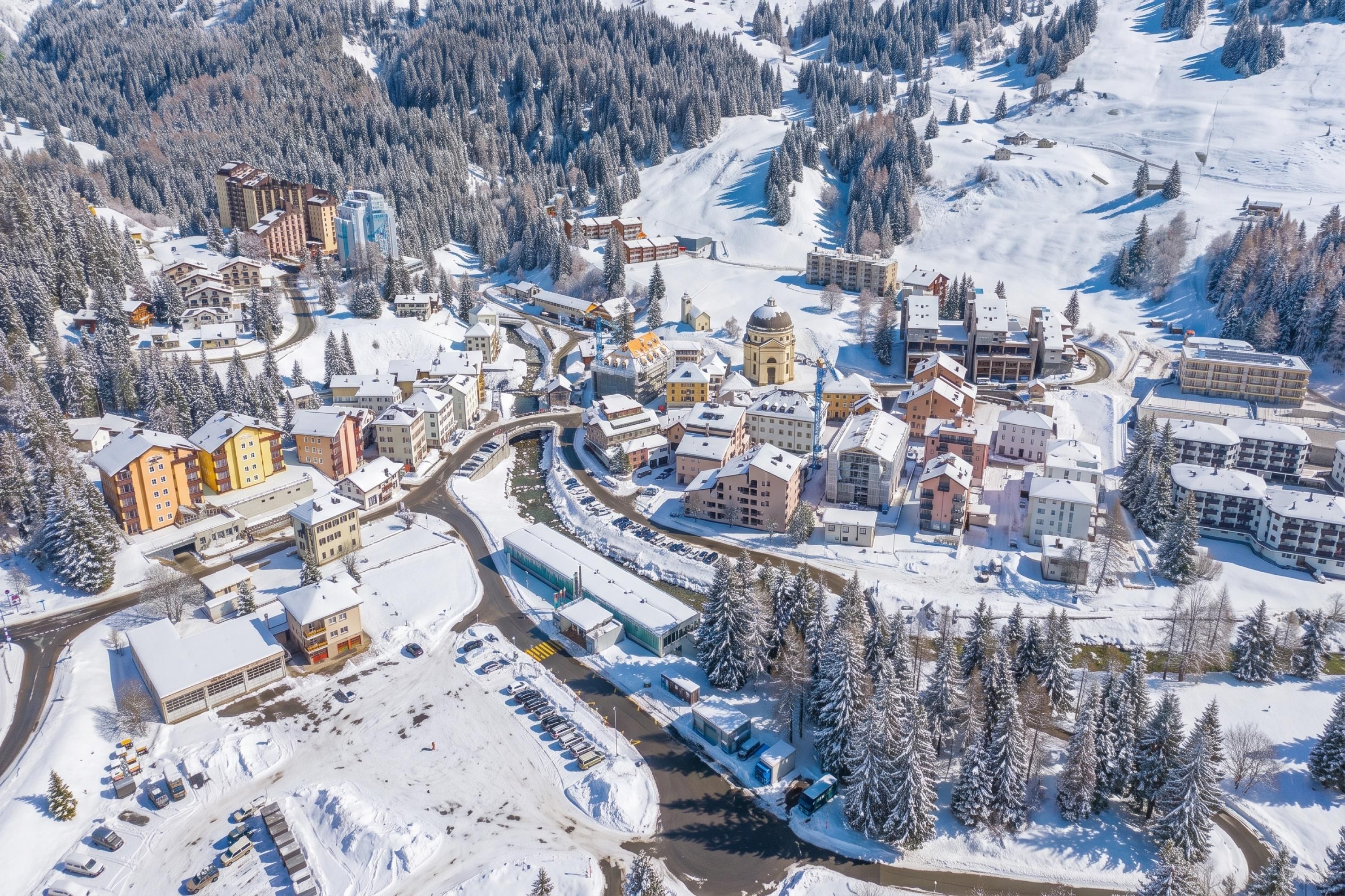 Eine verschneite Stadt mit einer Mischung aus modernen und traditionellen Gebäuden, einem Fluss, der hindurchführt, und einer bergigen Kulisse.