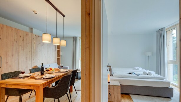 Una habitación de hotel moderna con zona de comedor de madera, cama con vistas y un ventanal grande.