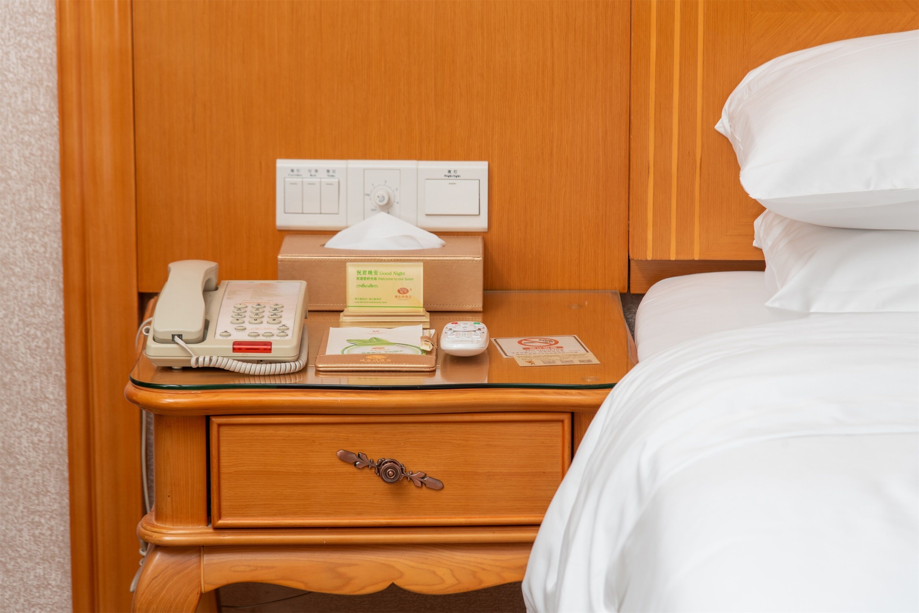 Una camera d'albergo con un comodino in legno, un telefono, una scatola di fazzoletti, un telecomando e un letto con biancheria da letto bianca.