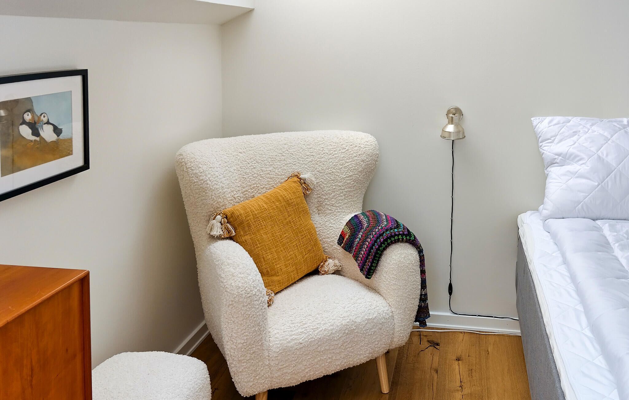 A room with a white armchair, a yellow pillow, a colorful knitted throw, a floor lamp, and a bed with white bedding.