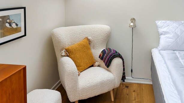 A room with a white armchair, a yellow pillow, a colorful knitted throw, a floor lamp, and a bed with white bedding. - 3 bedroom nice home in Tranekær (Tranekær)