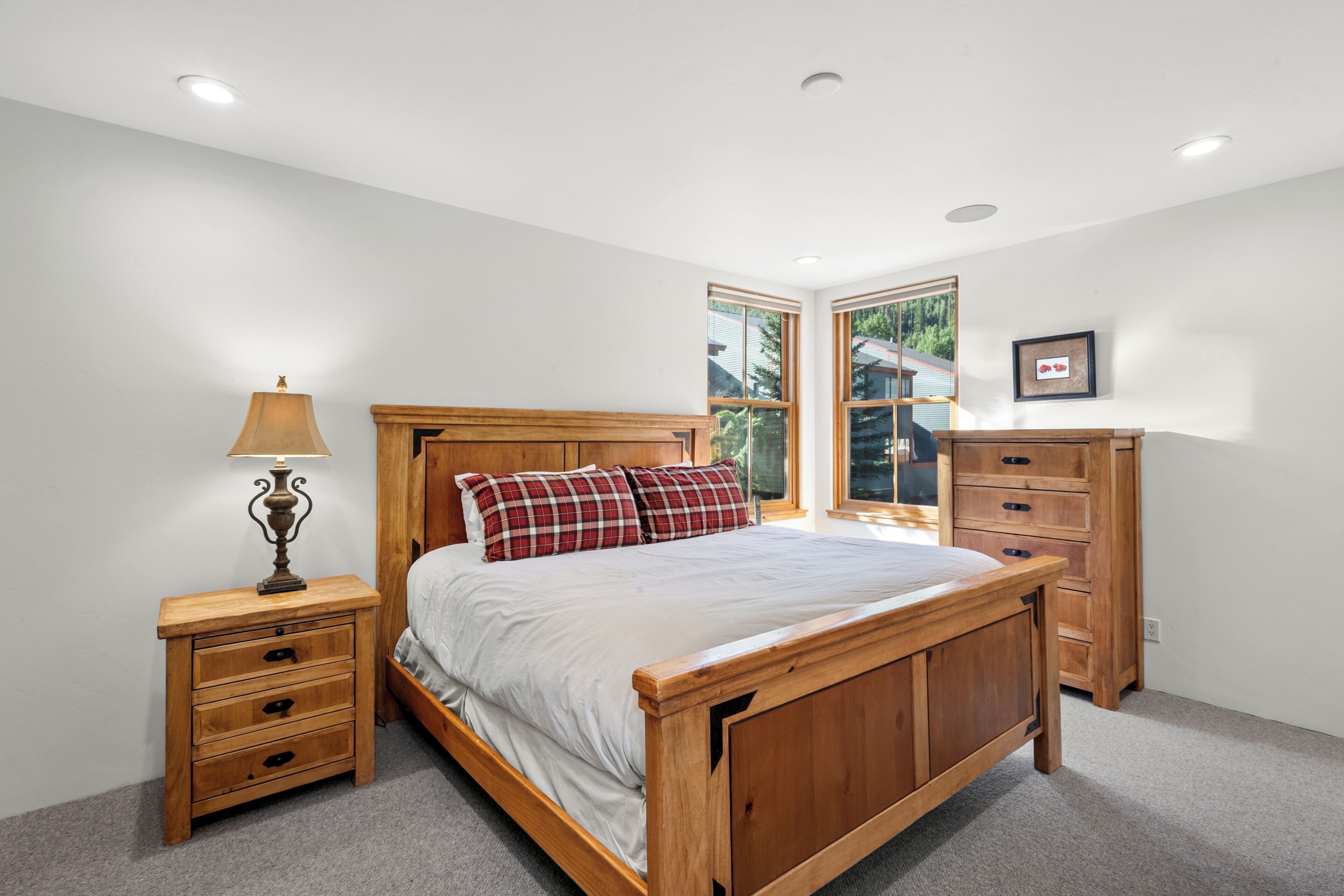 A bedroom with a wooden bed, a nightstand with a lamp, a dresser, and a window with a view of trees.