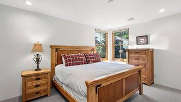 A bedroom with a wooden bed, a nightstand with a lamp, a dresser, and a window with a view of trees.