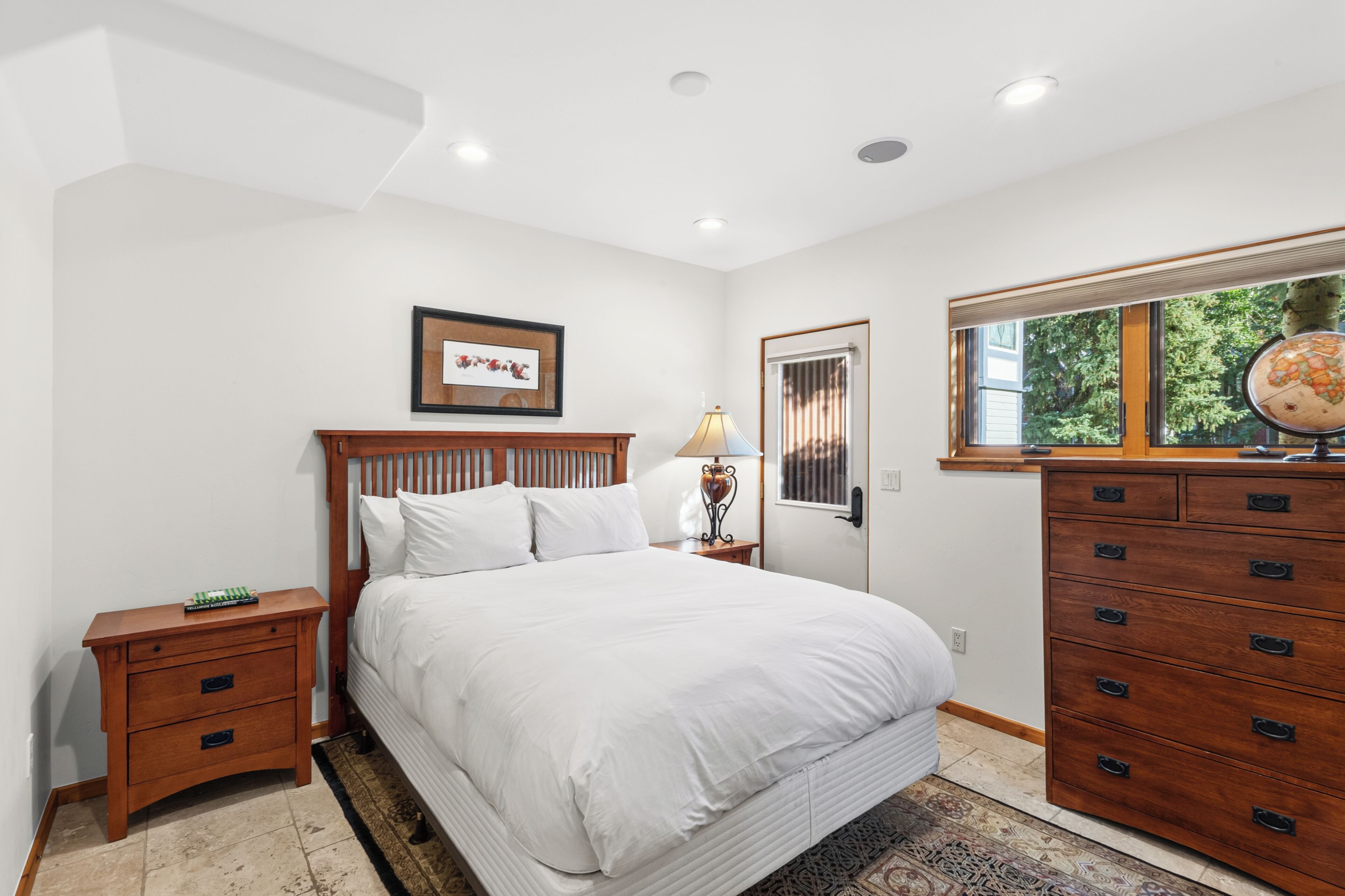 A bedroom with a large bed, a wooden dresser, a nightstand, a lamp, a framed picture on the wall, and a window with a view of trees.