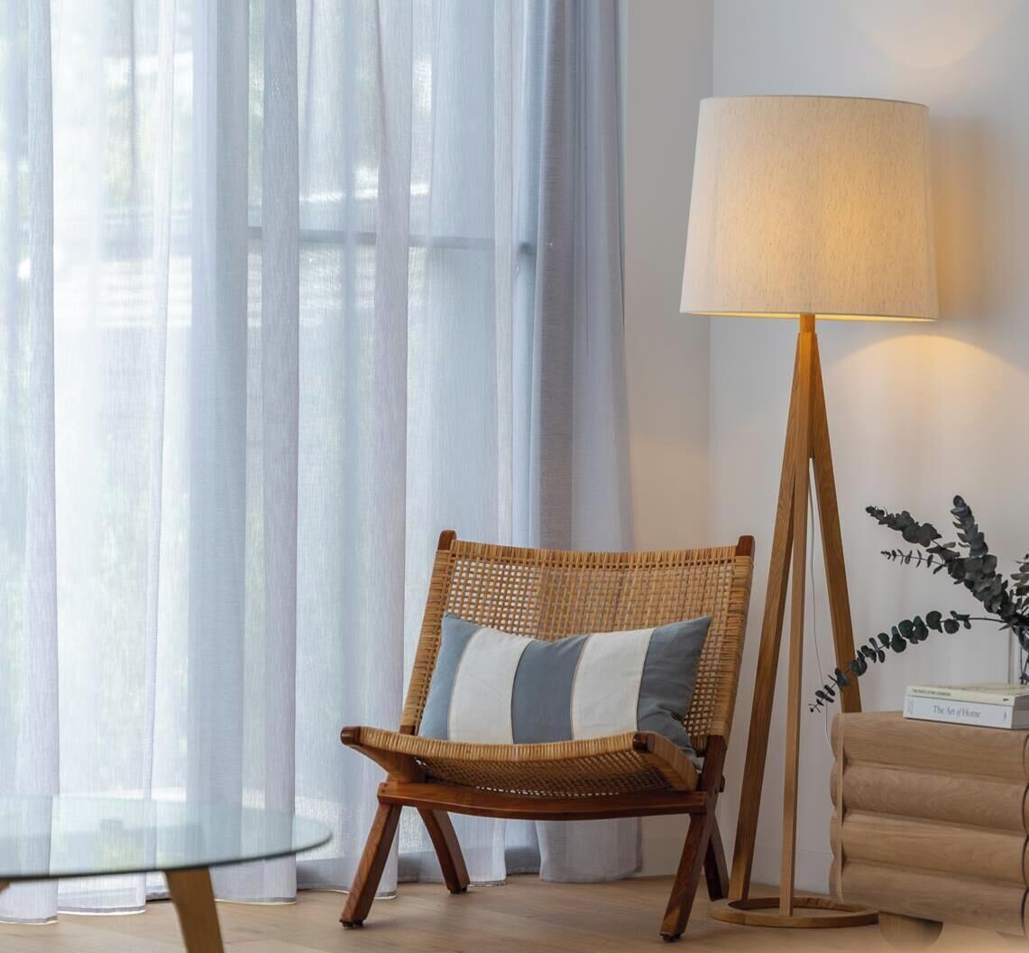 Une chaise en osier avec des coussins, un lampadaire et une table en verre, dans une pièce où sont accrochés de fins rideaux.