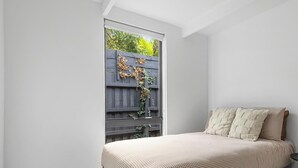 Ein Schlafzimmer mit grauem Bett, karierter Bettwäsche und Blick auf einen Holzzaun mit Grünpflanzen.