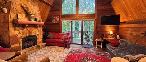 A cozy cabin interior with a stone fireplace, wooden walls, a red sofa, and a large window overlooking a forest.