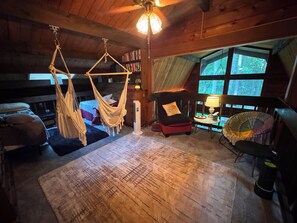 Um quarto aconchegante com uma rede, uma cadeira com almofada, uma mesinha de centro com abajur e uma janela grande com vista para árvores.