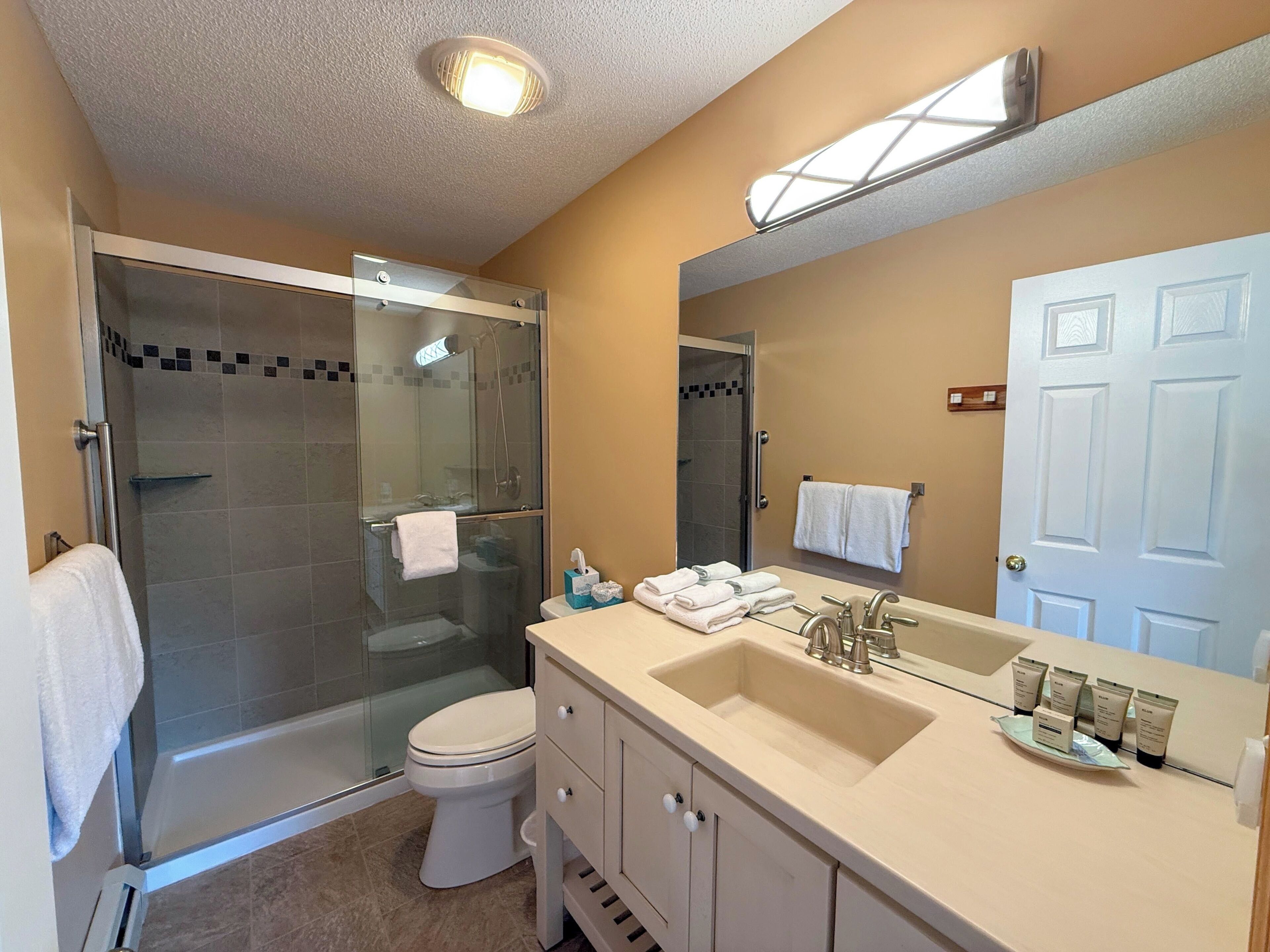 A bathroom with a glass-enclosed shower, a toilet, a sink with toiletries, and a mirror.