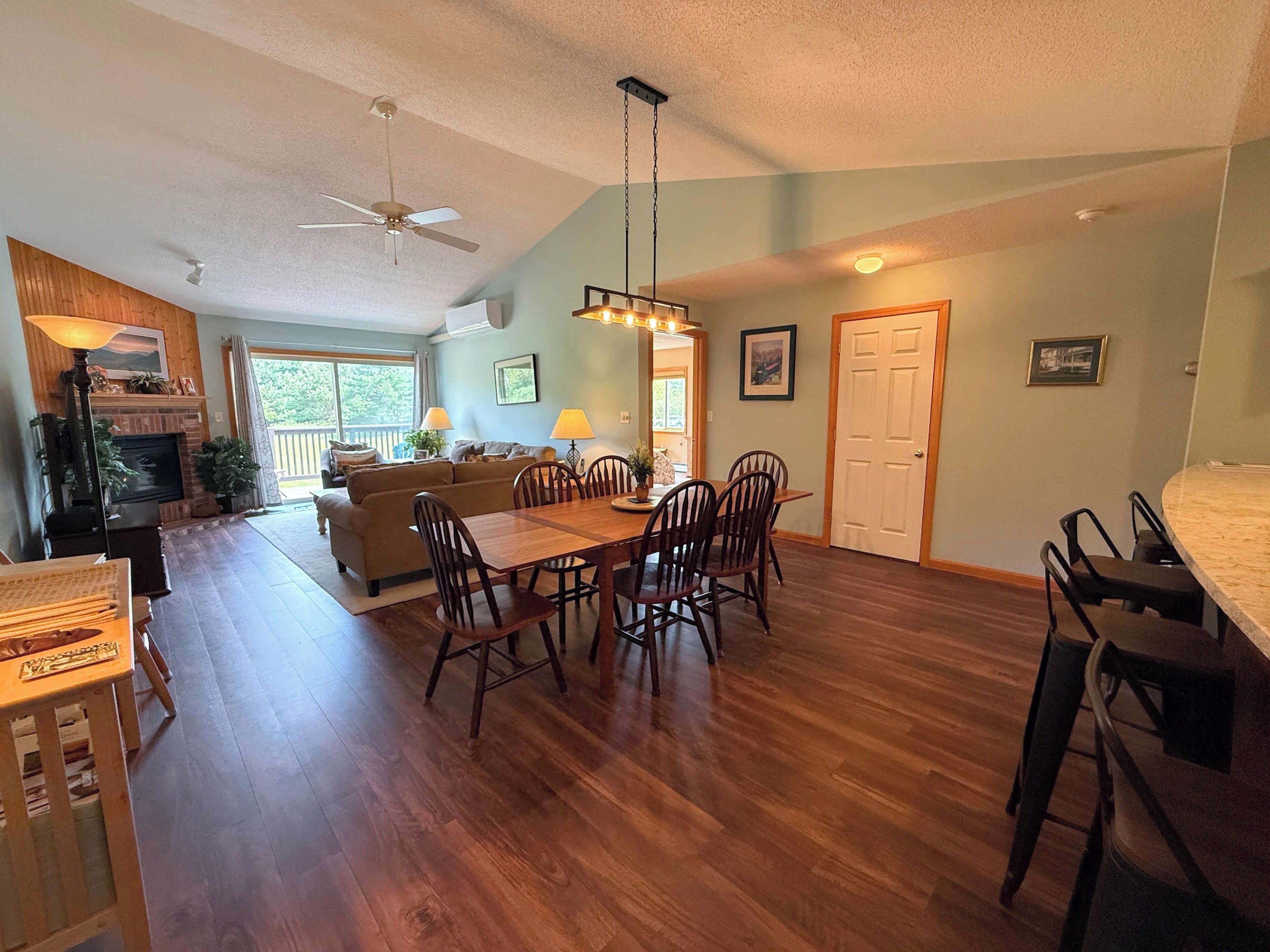 A spacious living area with a fireplace, dining table, and chairs. There is a ceiling fan, a large window, and a door leading to another room.