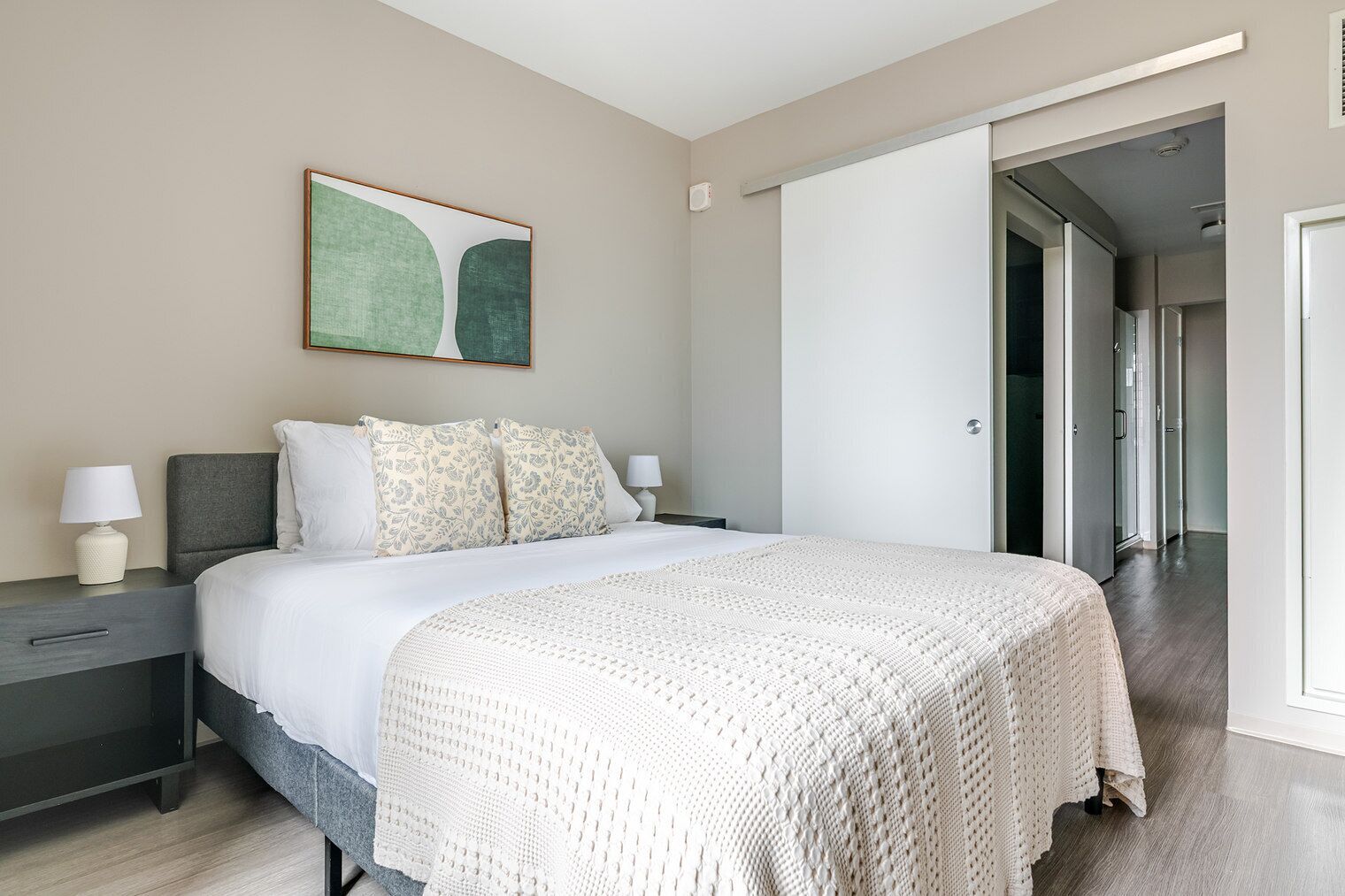 A modern bedroom with a bed, nightstands, a large window, and a door leading to another room.