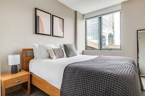 Une chambre d’hôtel avec un lit, une table de chevet, une lampe, deux tableaux encadrés, une fenêtre donnant sur la ville et un miroir.