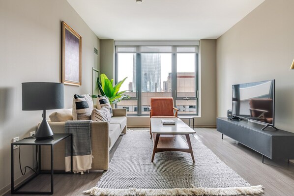 Un salon moderne comprenant un canapé, un fauteuil, une table basse, un téléviseur et une grande fenêtre donnant sur la ville.