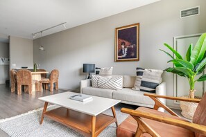 Un salon moderne avec un canapé, une table basse et des fauteuils. On y trouve également un coin repas avec une table et des chaises. Un tableau est accroché au mur, et une plante est placée dans un coin.