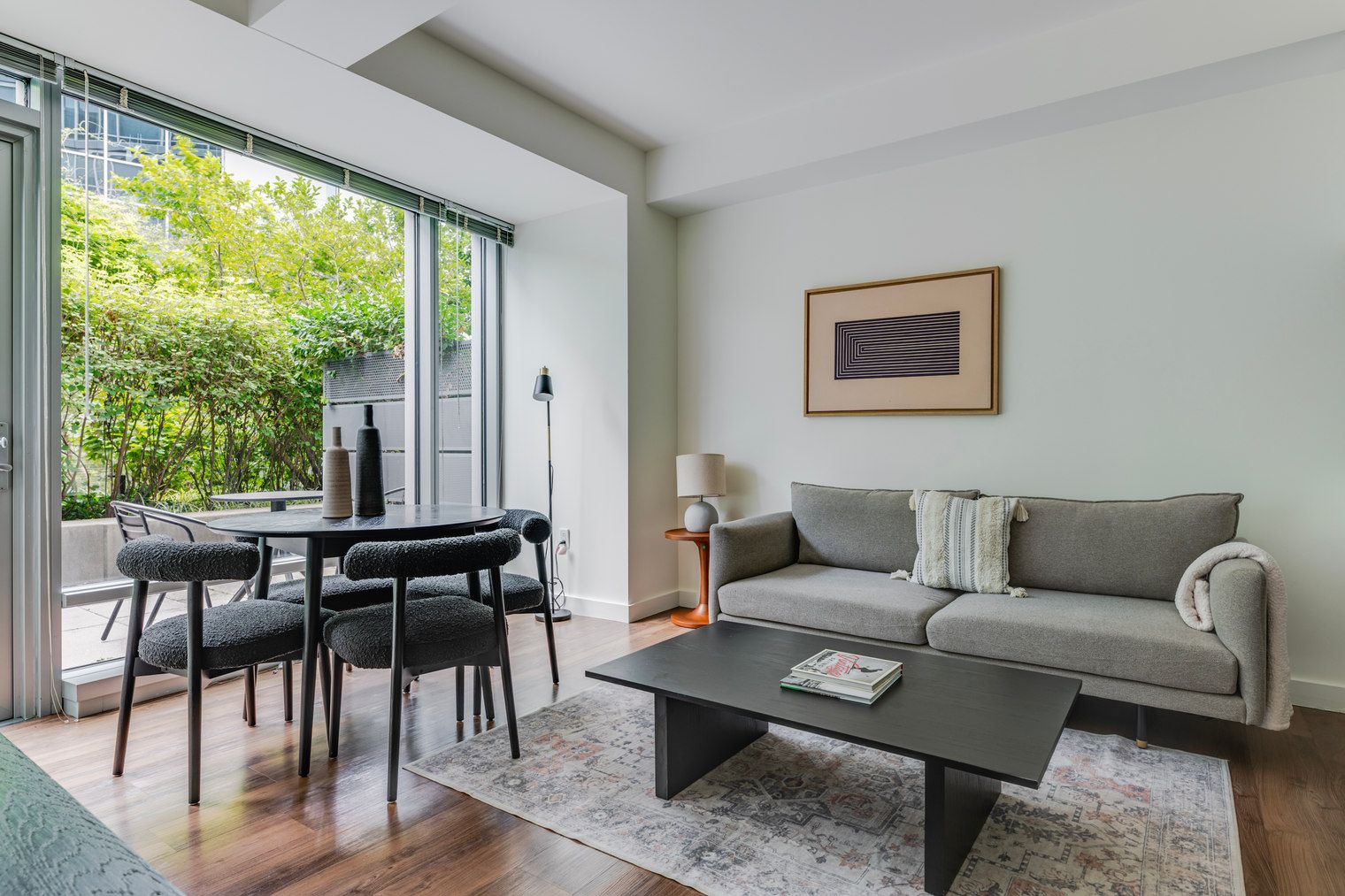A modern living room with a sofa, coffee table, and chairs, a dining area with a table and chairs, and a large window with a view of greenery.