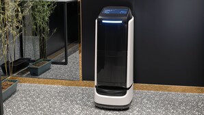 Un chiosco self-service con schermo digitale, su un pavimento maculato con una parete nera e una pianta sullo sfondo.