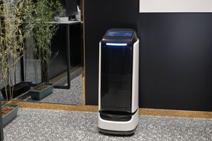 Ein Selbstbedienungskiosk mit digitalem Bildschirm, platziert auf einem gefleckten Boden vor einer schwarzen Wand und einer Pflanze im Hintergrund.