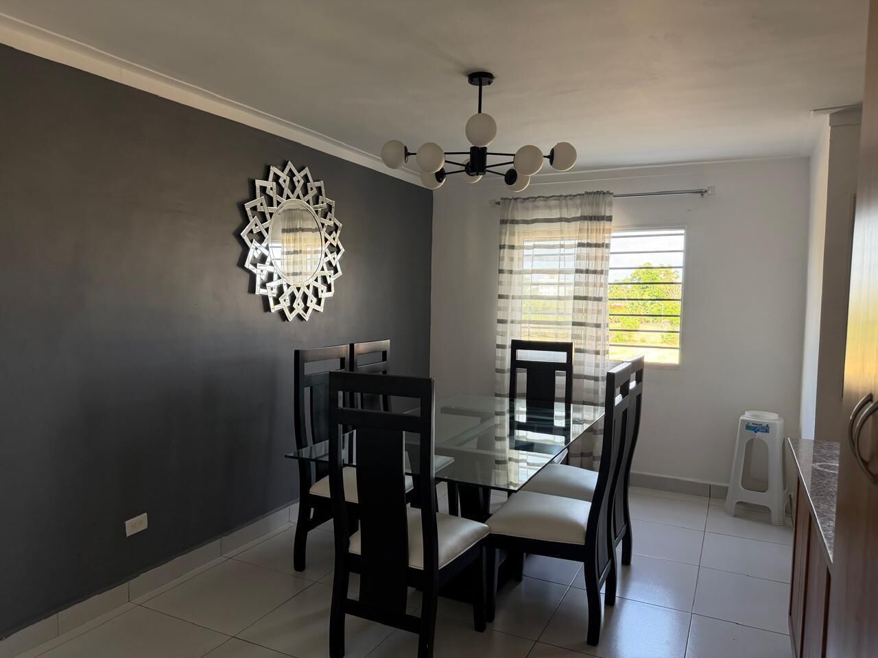 A dining area with a glass table and chairs, a decorative wall piece, and a window with curtains.