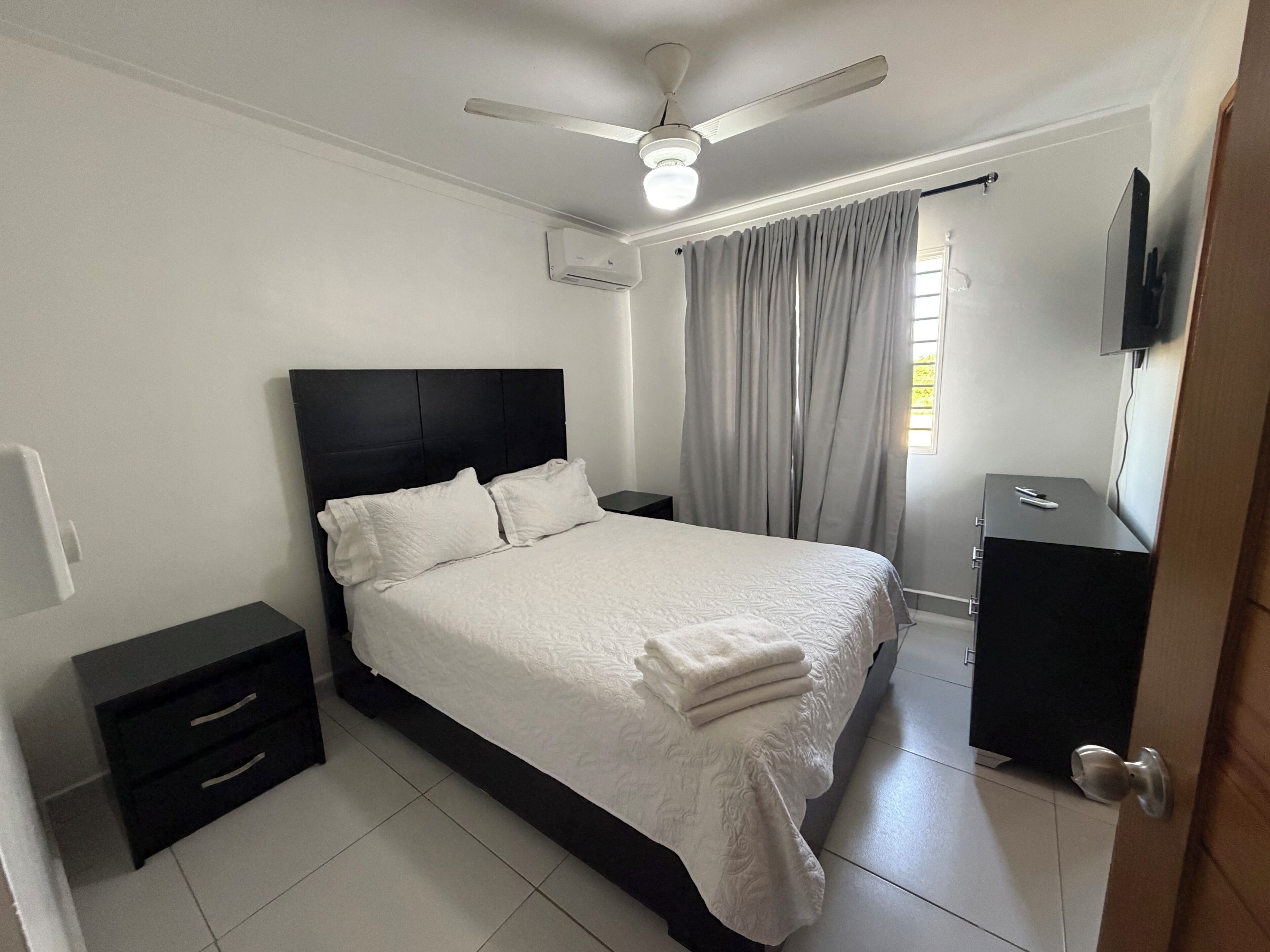 Un dormitorio con cama, mesita de noche, televisor, ventilador de techo y ventana con cortinas.