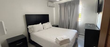 Un dormitorio con cama, mesita de noche, televisor, ventilador de techo y ventana con cortinas.