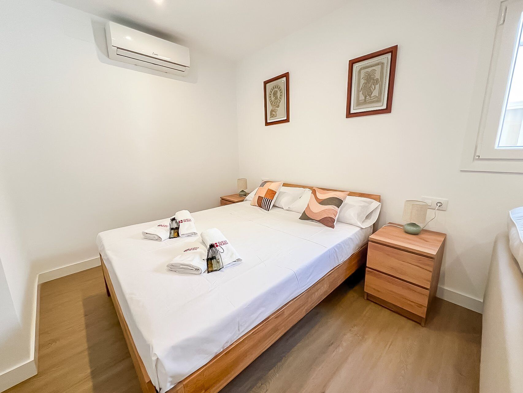 A hotel room with a single bed, a wooden nightstand, a bedside table, and an air conditioning unit on the wall.