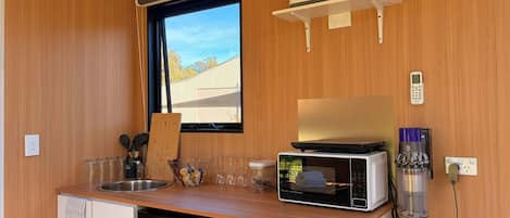 A compact kitchenette with a microwave, kettle, and a small shelf with a kettle and a cup.